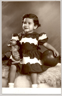 [Niñita de vestido rojo y blanco, con una pelota y un pato de juguete en sus manos mirando hacia el lado]