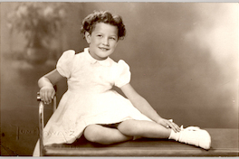 [Niña rubia de ojos claros y pelo corto ondulado sentada con sus piernas cruzadas, vestido mangas cortas y zapatos y soquetes blancos]