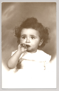 [Niñita de vestido blanco con vuelos pelo crespo ojos grandes y un dedo metido en la boca]