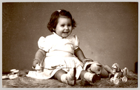 [Niñita sonriente de vestido blanco con mangas englobadas, calcetines y chalas blancas, sentada sobre un felpudo con varios juguetes]