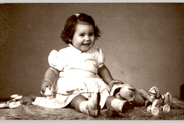 [Niñita sonriente de vestido blanco con mangas englobadas, calcetines y chalas blancas, sentada sobre un felpudo con varios juguetes]