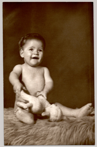 [Bebé desnudo sentado sobre un felpudo con un juguete sobre sus piernas]