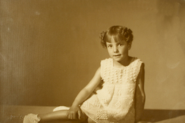 [Niñita rubia de vestido color blanco de hilo grueso y zaptillas blancas, sentada con las piernas hacia el lado]