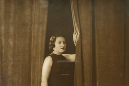 [Mujer joven y regordeta de pie con un vestido largo posando afirmada entre dos cortinas]