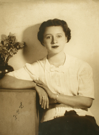 [Mujer joven sentada y apoyada en un mueble mirando de frente con una blusa blanca y falda negra]