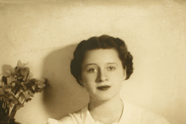 [Mujer joven sentada y apoyada en un mueble mirando de frente con una blusa blanca y falda negra]