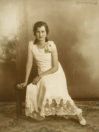 [Adolescente de vestido blanco largo adornado con broderí en la parte inferior sentada en un piso de madera sobre una alfombra que cubre la sala]