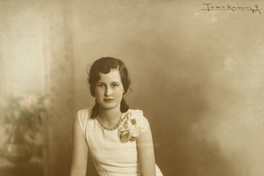 [Adolescente de vestido blanco largo adornado con broderí en la parte inferior sentada en un piso de madera sobre una alfombra que cubre la sala]