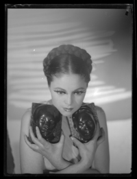 [Mujer joven, peinada con una trenza alrededor de su cabeza, sus manos cruzadas sobre el pecho, sostienen dos pequeños rostros esculpidos en bronce]