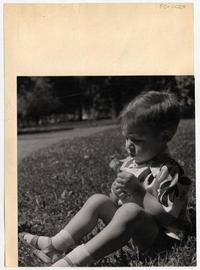 [Pequeño niño, con una flor en sus manos, sentado en la hierba del parque]