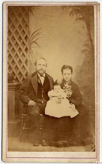 [Padres sentados en un sillón de madera y una bebé en la falda de la madre]