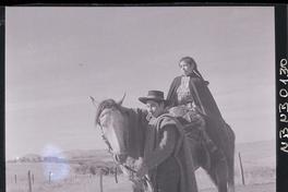 [Mujer mapuche a caballo, su marido tira las riendas por un camino rural]