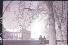 [Una pareja paseando por el Parque Forestal, en la ladera opuesta del río Mapocho, la Escuela de Derecho de la Universidad de Chile]