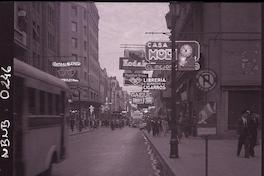 [Transeúntes por una calle con avisos luminosos de casas comerciales, como Musalem Hnos., Central Optica Hammersley Hnos, Kodak y otras]