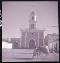 [Niña con un burrito que lleva carga, pasa frente a una capilla]