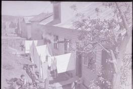 [Tres niños pequeños y una mujer que tiende ropa junto a su vivienda en un sector popular]