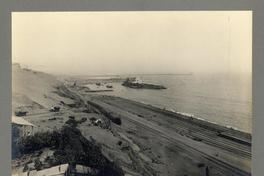 [Vista panorámica del puerto, se divisan vías férreas a orillas del mar, unas casas de la faena, y barcos en el mar]