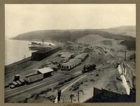 [Vista panorámica del puerto, se divisa el muelle, terminal ferroviario con una locomotora, vías férreas y almacenes]