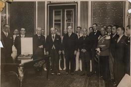 [Retrato de Manuel Rivas Vicuña, junto a un grupo de personas, en una ceremonia de condecoración; En una mesita, se encuentra la caja que guarda la distinción]