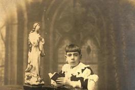 [Retrato de un niño en el día de su Primera Comunión, en sus manos sostiene un misal y rosario, se encuentra en un reclinatorio, junto a una mesita con la imagen de la Virgen]