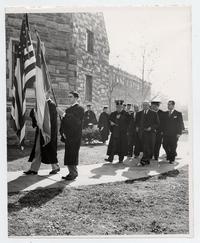 [Guillermo Feliú Cruz junto a otros señores en ceremonia de graduación, caminando por costado de edificio, detrás de los estandartes chileno y norteamericano. Estados Unidos]