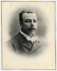 [Retrato plano medio corto de un hombre joven, de cabello corto peinado hacia atrás y bigote, viste traje, camisa de cuello alto y corbata-humita]