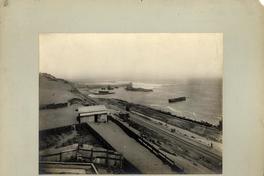 [Vista panorámica del puerto, se divisan vías férreas a orillas del mar, unas casas de la faena, y barcos en el mar]