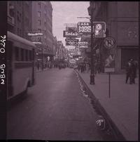 [Transeúntes por una calle con avisos luminosos de casas comerciales, como Musalem Hnos., Central Optica Hammersley Hnos, Kodak y otras]