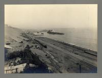 [Vista panorámica del puerto, se divisan vías férreas a orillas del mar, unas casas de la faena, y barcos en el mar]