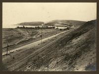 [Vista panorámica del puerto, desde el cerro, se divisa el muelle, almacenes, vías férreas y unos barcos en el mar]